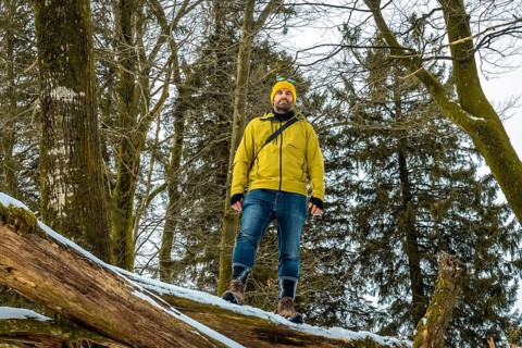 Man in yellow jacket and beanie standing on a fallen log in a snowy forest, confidently taking in a
