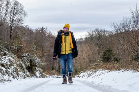 Man in warm winter attire and sunglasses walking on a snowy road through a woodland landscape,