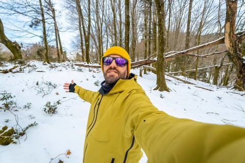 Man posing for a selfie during a winter hike, exploring a snowy forest landscape with bare trees