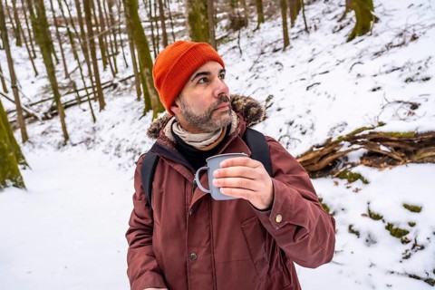 Man wearing an orange beanie and winter jacket, holding a hot mug while contemplating the serene