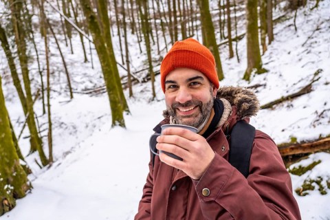 Bearded man smiling in a winter hat and jacket, holding a steaming mug while standing in a snow