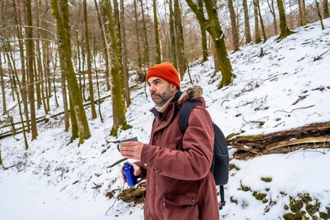 Adventurous man in a beanie and jacket pauses on a snowy forest trail with a backpack, sipping a