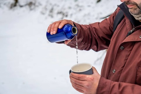 Man wearing a winter jacket pouring a hot drink from a blue thermos bottle into a cup, staying