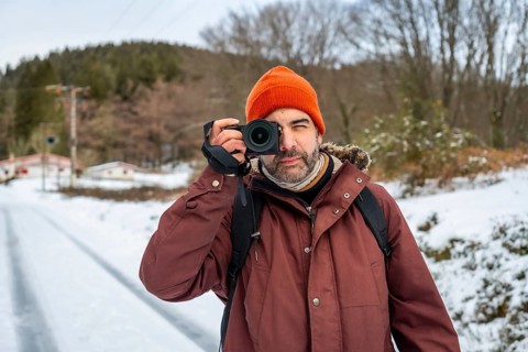 Man in winter clothing and an orange beanie holding a mirrorless camera, capturing nature and