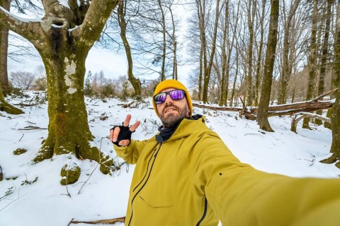 Man smiling at the camera and making a peace sign gesture, wearing a yellow jacket, beanie, and