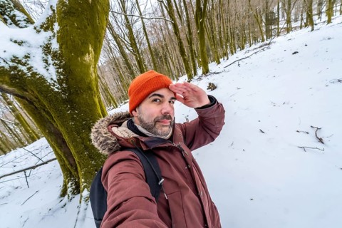 Man posing for a selfie in a snowy forest during winter, looking out with his hand shading his