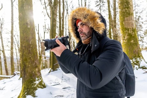 Man in winter parka with fur hood photographing a snowy forest trail, capturing nature with a large