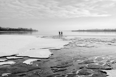 Two people walking on a frozen lake under a cloudy sky in a wintry landscape, black and white