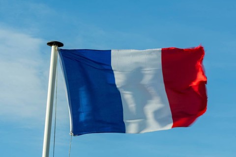 A French flag flutters in the wind on a white flagpole. The background is a clear, azure sky