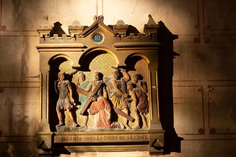 A carved relief depicts a religious scene. Jesus falls while carrying the cross. Roman soldiers and