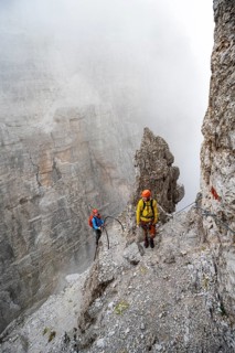 Two mountaineers climb the Via Ferrata Oliva Detassis via ferrata in fog, steep mountains covered
