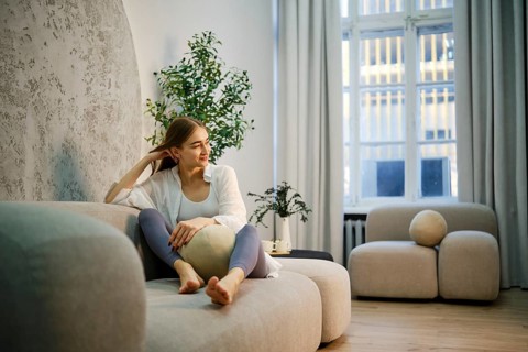 A cute woman is sitting on a light sofa in a modern room. She has a relaxed posture and is holding