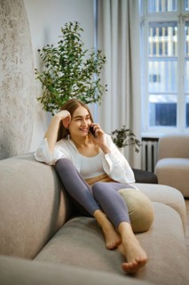 Barefoot woman is seated on a couch in a room with soft lighting. She is smiling while talking on
