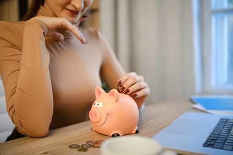 A woman sits at a wooden desk, placing coins into a pink piggy bank. She is focused on saving money