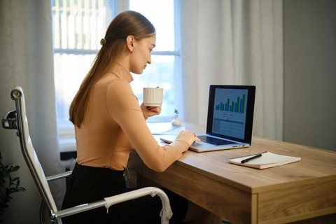 A woman sits at a wooden desk while using a laptop. She holds a cup in one hand and looks at the