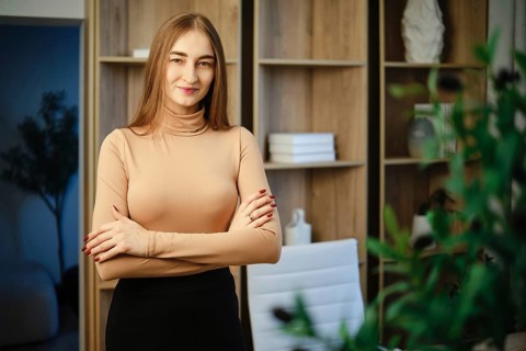 A woman with long hair stands in a modern room with wooden shelves. She has her arms crossed and