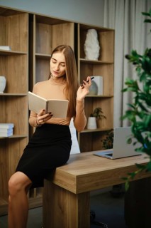 A woman sits on a wooden desk in an office. She is writing in a notebook while holding a pen. A