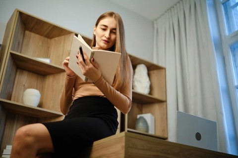 A woman sits on a wooden desk with a notebook in hand. She is focused on reading while surrounded