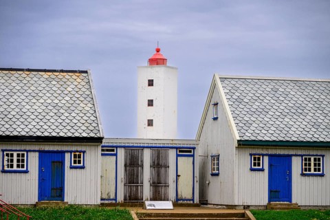 Lighthouse between two blue and white houses rises under a grey sky Kjølnes Fyr, small windows and