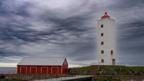 White lighthouse next to a red wooden building against a cloudy sky at the sea Kjølnes Fyr, small
