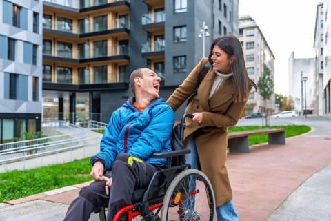 Man with a disability in a wheelchair laughing happily while a female caregiver pushes him along a