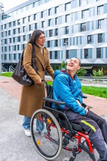 Caregiver pushing a man with a disability in a wheelchair outdoors, both smiling and sharing a