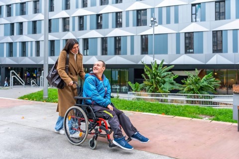 Happy woman caregiver pushing smiling adult man in a wheelchair on an accessible ramp in an urban