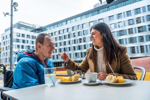 Woman feeding a smiling man with a disability, enjoying a meal and coffee at an outdoor cafe,