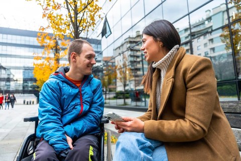 Man in wheelchair smiling and attentively listening to a woman sitting beside him on a bench,