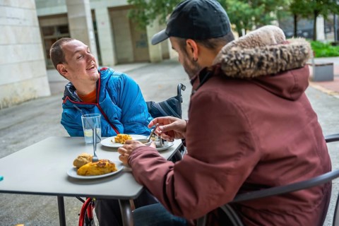 Two friends sharing an outdoor meal on a city terrace, both men are using wheelchairs and laughing,