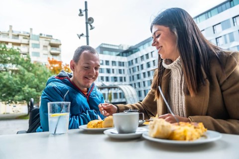 Smiling caregiver offering food to a man in a wheelchair, enjoying a pleasant conversation and meal