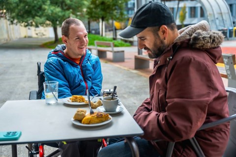 Young man with a disability smiling and relaxing in a wheelchair at an outdoor cafe table, enjoying