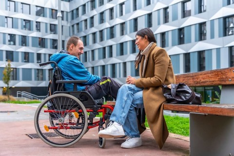 Man in a wheelchair and woman sitting on a bench engaging in a friendly conversation outdoors,