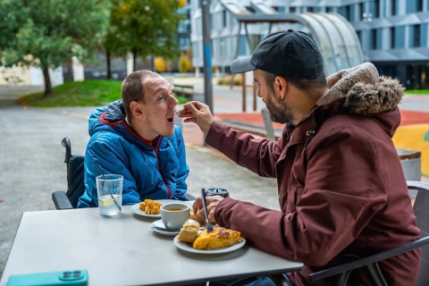 Man tenderly feeding a young boy in a wheelchair at an outdoor cafe, representing care, support,