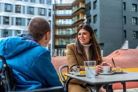 Woman and boy in a wheelchair share a warm, engaged conversation at an outdoor urban cafe, smiling