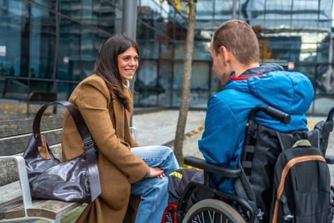 Woman sitting on bench chatting and laughing with a boy in a wheelchair in a city park, warm
