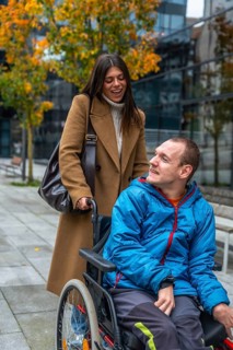 Cheerful caregiver standing behind a young man in a wheelchair, pushing him along an urban sidewalk