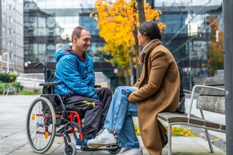Young man living with disability in a wheelchair smiling and talking with a woman sitting on a park