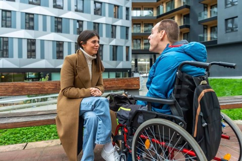 Woman and man with cerebral palsy sitting together by a bench, having a conversation in an urban