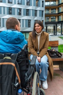 Young woman sitting on a park bench talking and smiling with a man in a wheelchair, showing