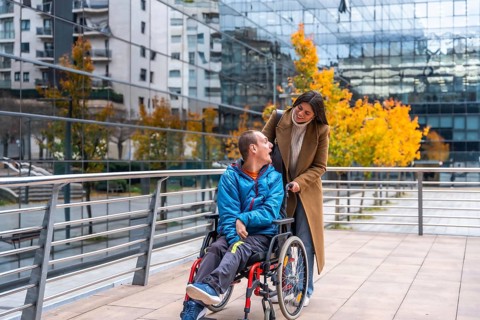 Caregiver pushing an adult man in a wheelchair on a modern city street, both individuals smiling