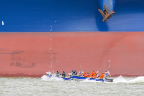 THW lifeboat with crew in orange clothing sails in front of a large ship hull on rough water,