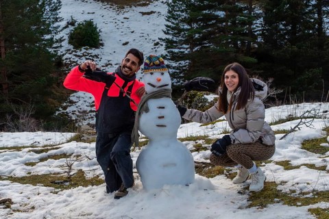 Couple building a snowman together on a snowy mountain slope, smiling and bundled in winter gear,