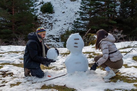 Young couple building a snowman on a sunny mountain slope, laughing and bonding in warm winter