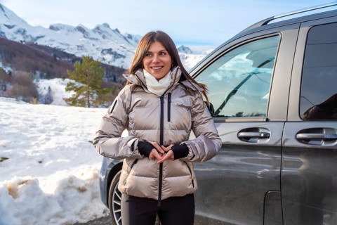 Woman standing by a vehicle against snowy mountain peaks, wearing a warm jacket and scarf, smiling