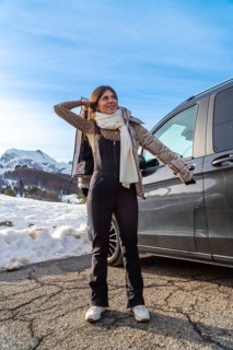 Young woman standing on a snowy mountain road next to a dark vehicle, wearing winter clothes