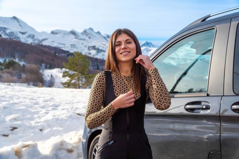 Young woman smiling while standing next to a car, enjoying a winter road trip adventure with snow