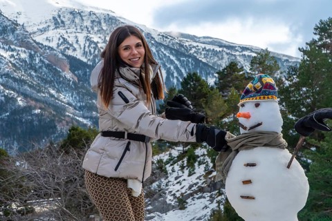 Young woman smiling and wearing winter clothing, building a snowman with a colorful hat and scarf