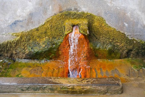 Rust-brown water flows from a spring in a concrete wall surrounded by eroded mineral deposits,