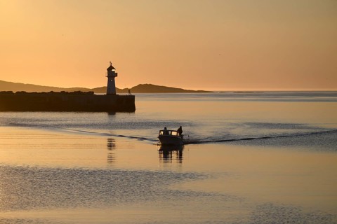 A small motorboat sails through the harbor entrance with lighthouse and midnight sun, Vardø,